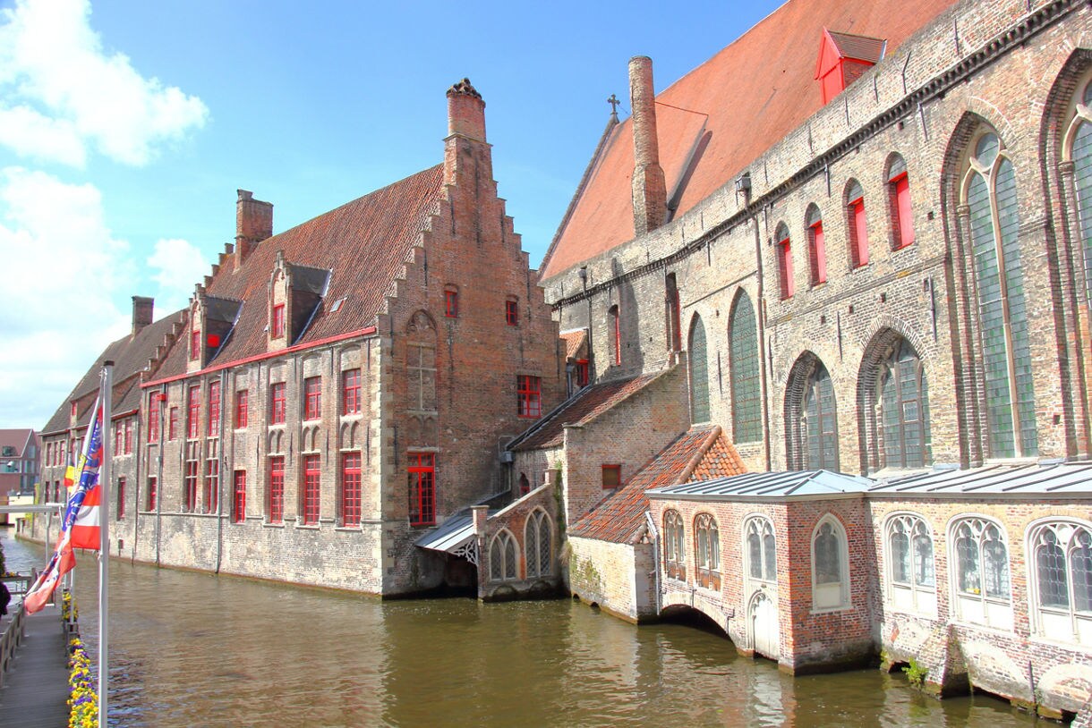Historic brick buildings with red windows lining a canal in Bruges, Belgium, featuring gabled roofs and arched windows reflected in the water.