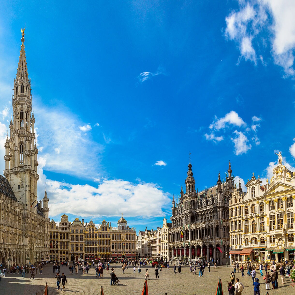 Panoramic view of the Grand Place in Brussels, Belgium, with the ornate Town Hall tower, historic guildhalls and bustling crowds under a bright blue sky.