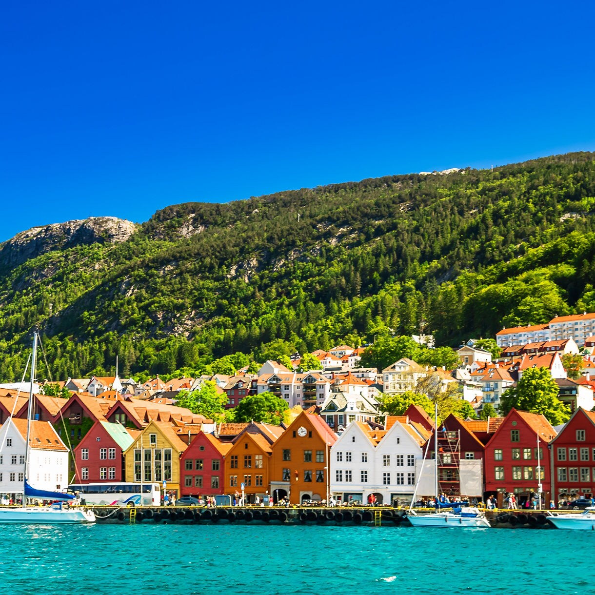 Brightly painted wooden buildings line Bryggen Wharf in Bergen, Norway, with forested hills rising in the background.