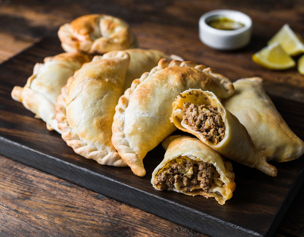 A pile of baked empanadas on a wooden board with one cut open to show seasoned meat inside.