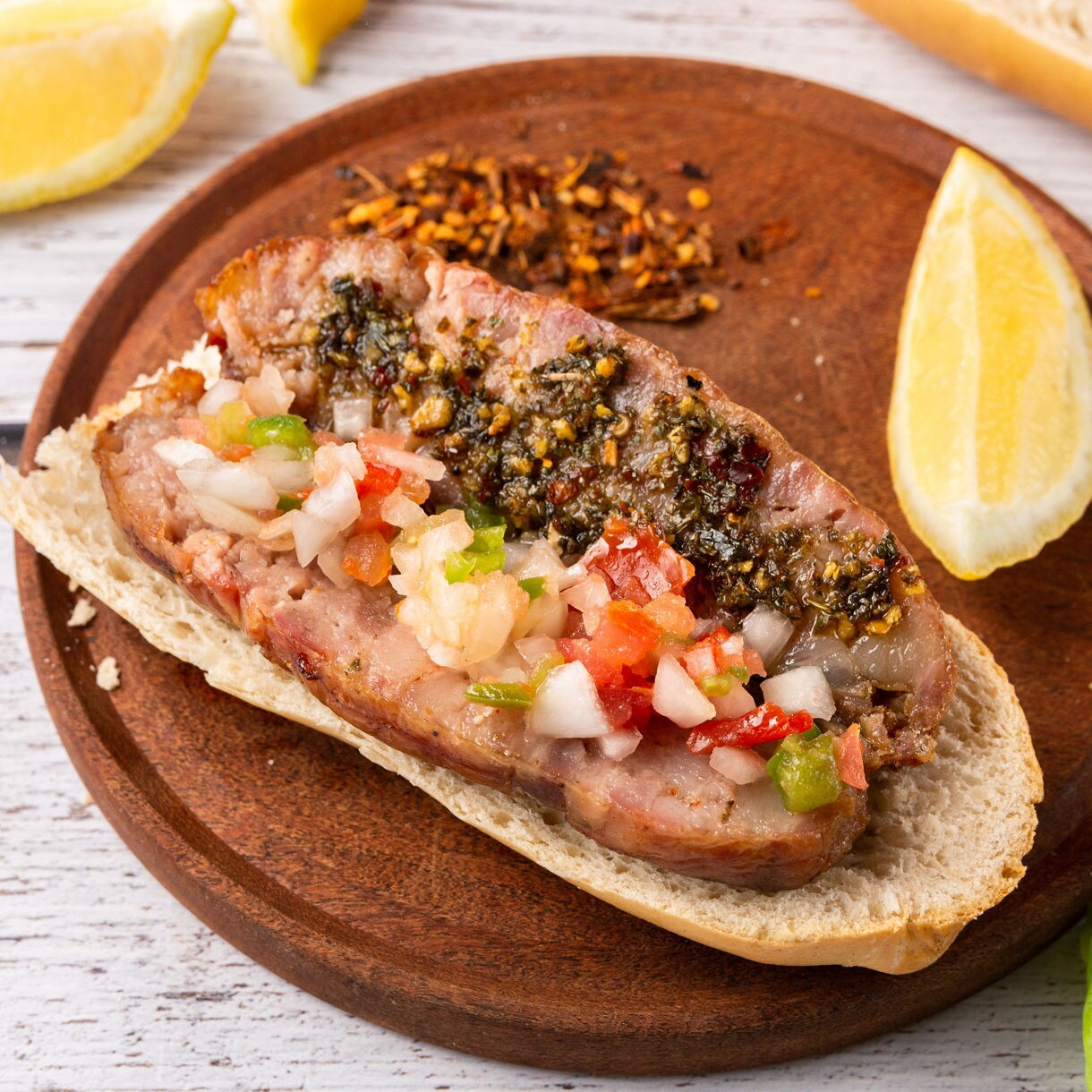 Grilled sausage in bread topped with chimichurri and diced vegetables, served with lemon wedges.