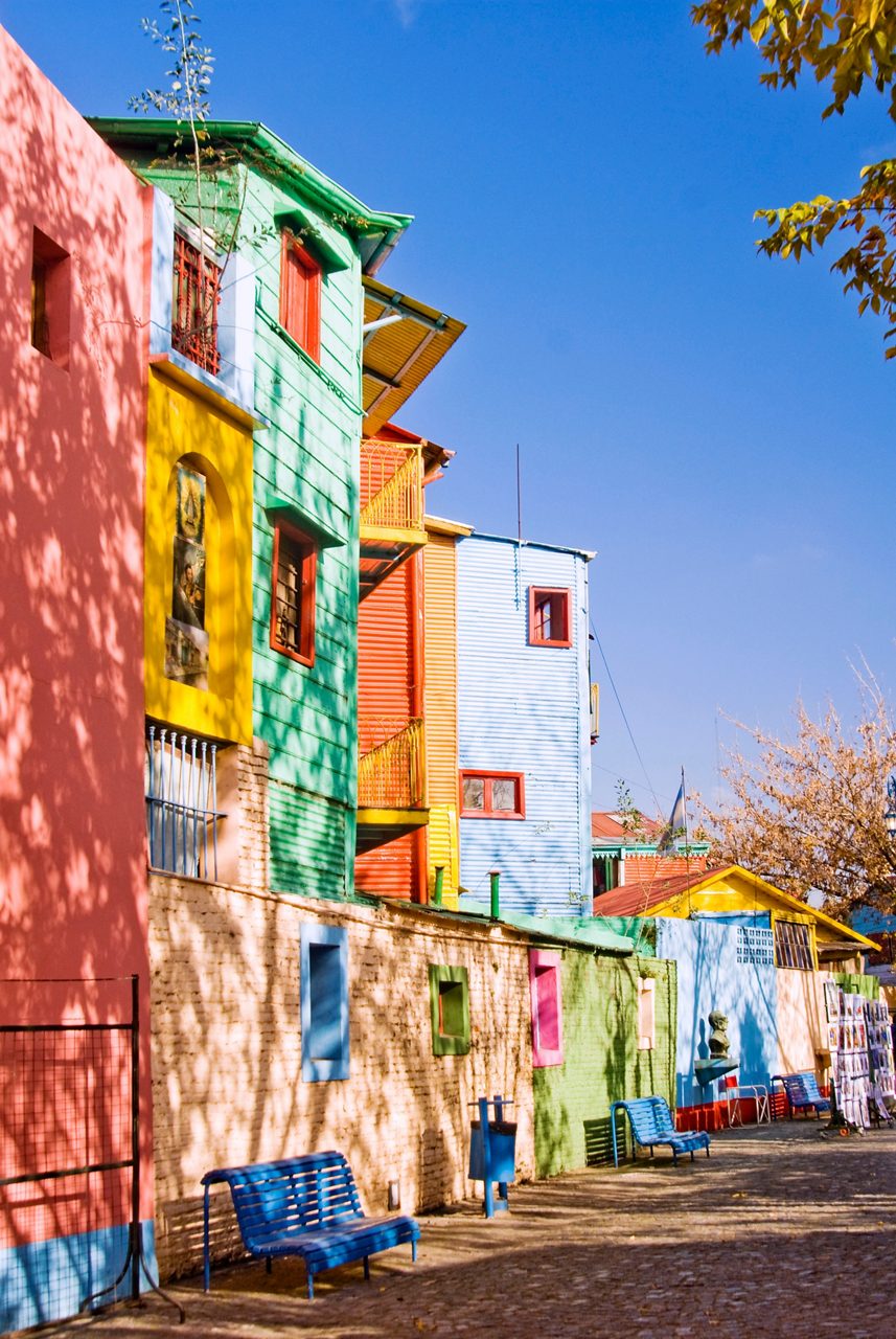 Vibrant houses in pink, green, yellow and blue line a cobblestone street with blue benches in the shade