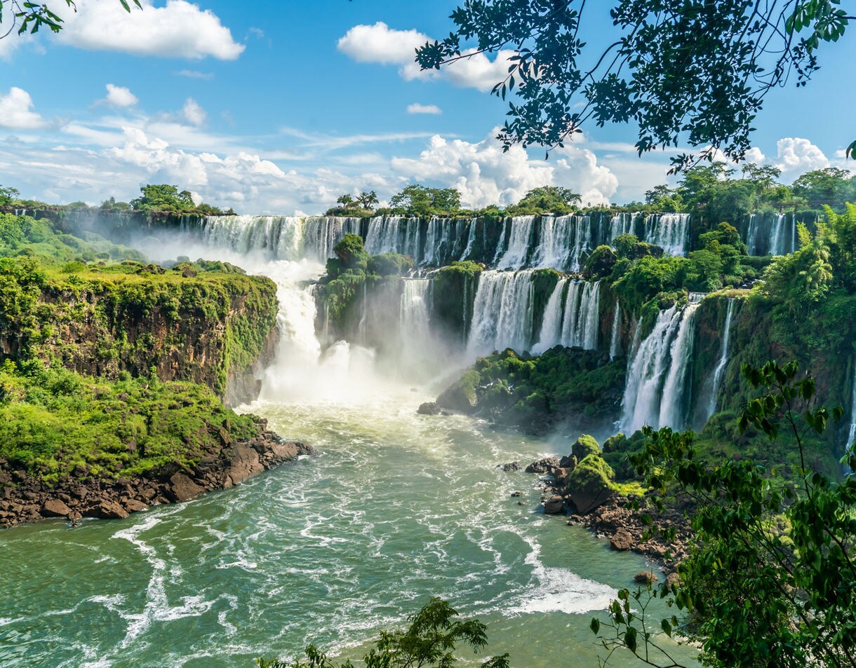 Wide view of Iguazú Falls with multiple waterfalls pouring over lush green cliffs into a misty basin.