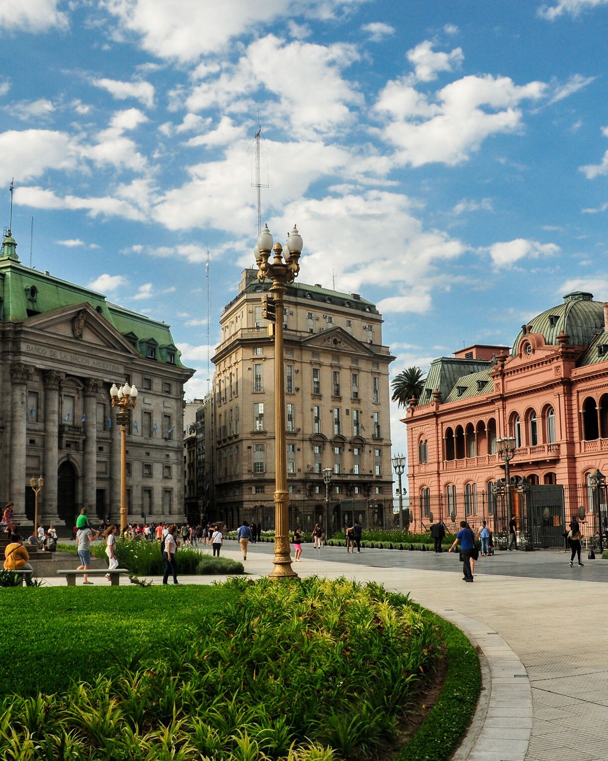 Wide plaza with green lawns surrounded by historic buildings including the pink Casa Rosada.