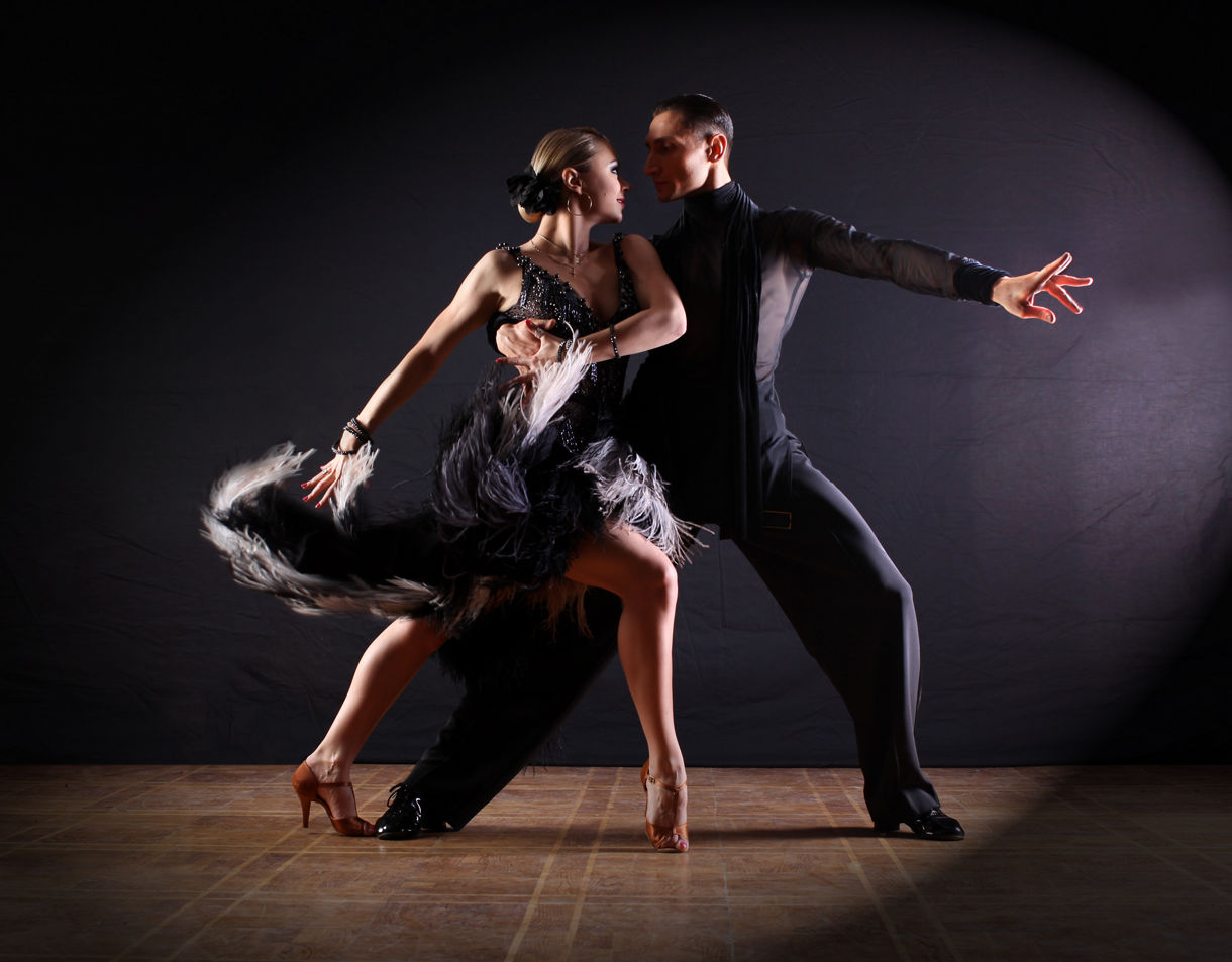 A couple performing a dramatic tango pose on a dimly lit stage with swirling fringe on the woman’s dress.