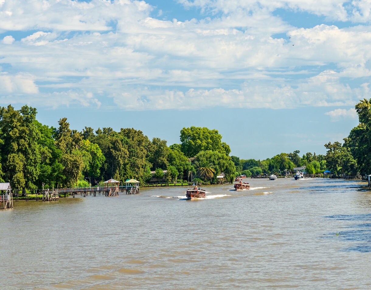 Boats speeding along a wide brown river bordered by dense green trees and small docks.