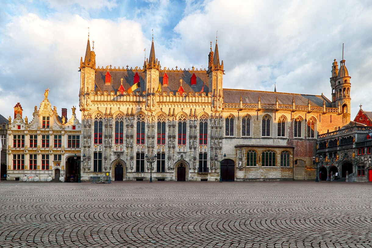Wide view of Burg Square in Bruges, Belgium, featuring the ornate Gothic City Hall with red-trimmed windows, gilded Renaissance buildings and a cobblestone plaza in the foreground.