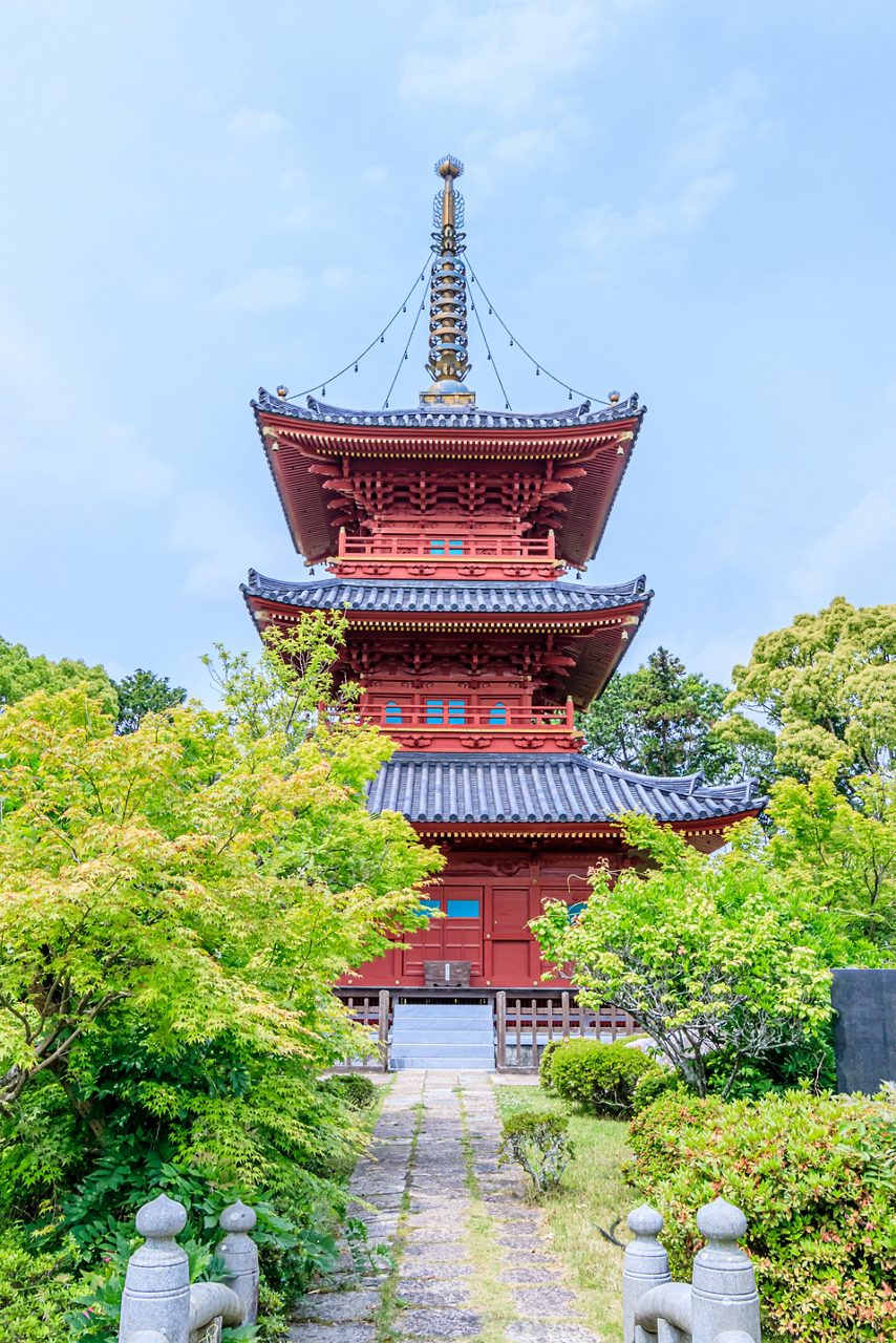 A vivid red three-story pagoda at Buzen Kokubunji Temple surrounded by lush green foliage and framed by a stone path leading to its entrance.