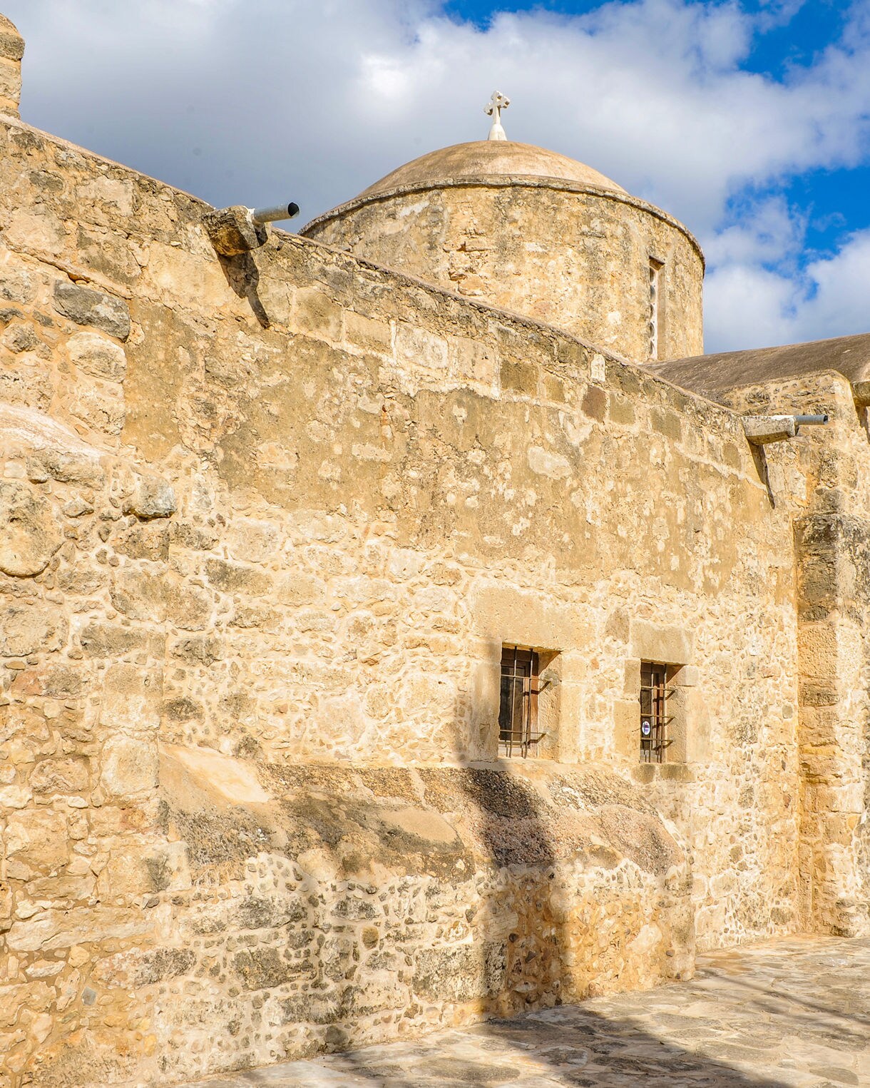  Ancient Byzantine church built of golden stone with an arched roof and small barred windows, framed by cypress trees and a bright blue sky in Rhodes, Greece.