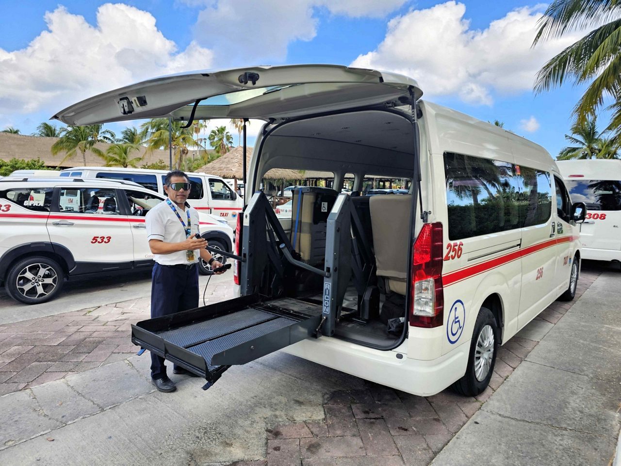 A crew member stands beside a wheelchair-accessible van with a lowered ramp deployed at a sunny port destination.