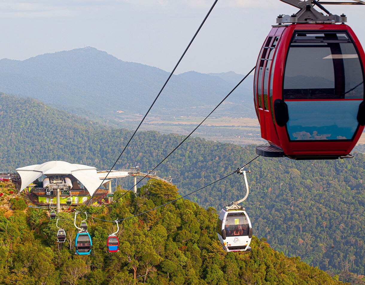 Colorful gondolas of the Langkawi SkyCab cable car gliding above dense green forest with mountain ranges in the distance.