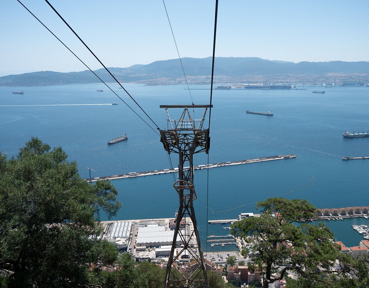 A cable car tower and cables descending toward Gibraltar’s harbor, with ships scattered across deep blue water and mountains in the distance.