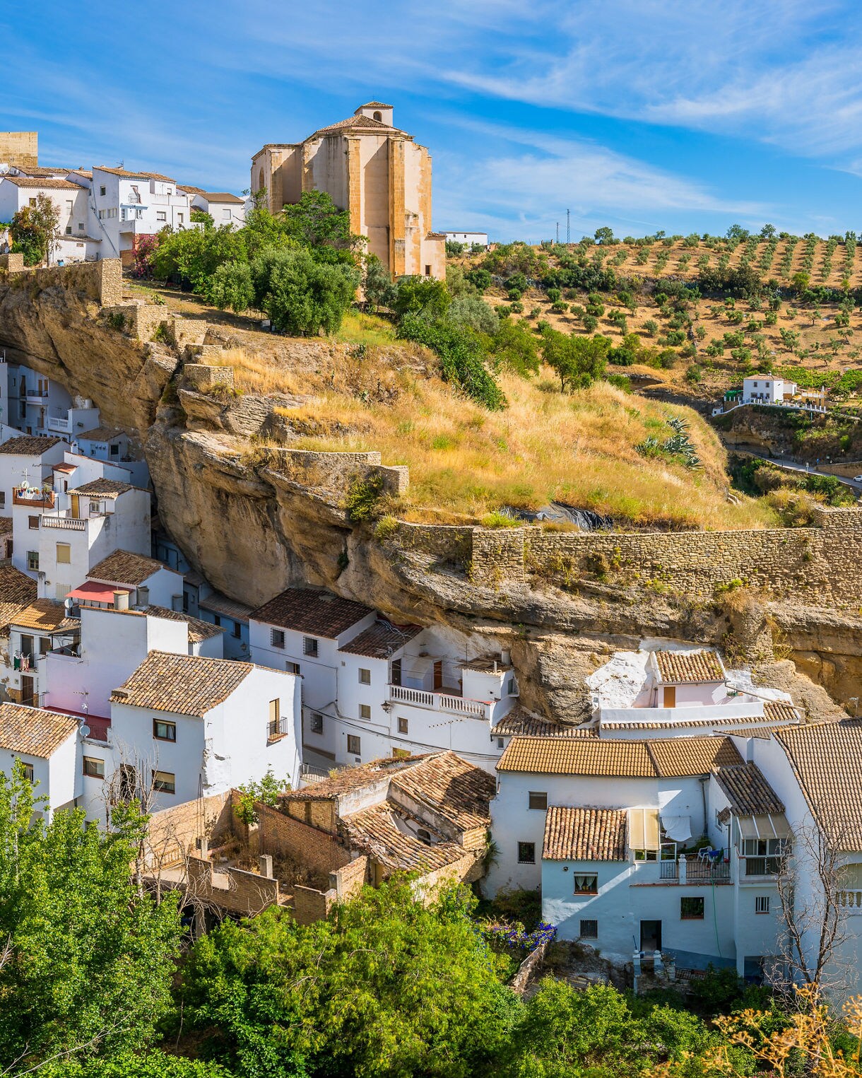 Hillside village of Setenil de las Bodegas in southern Spain, with white houses built directly into rocky cliffs and rolling fields in the background under a bright blue sky.