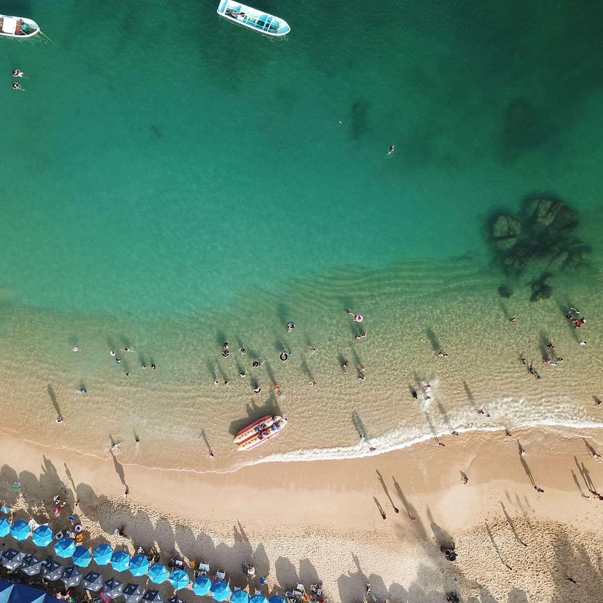 Aerial view of Caleta Beach in Acapulco with turquoise water, swimmers, anchored boats and rows of blue umbrellas on the sand.