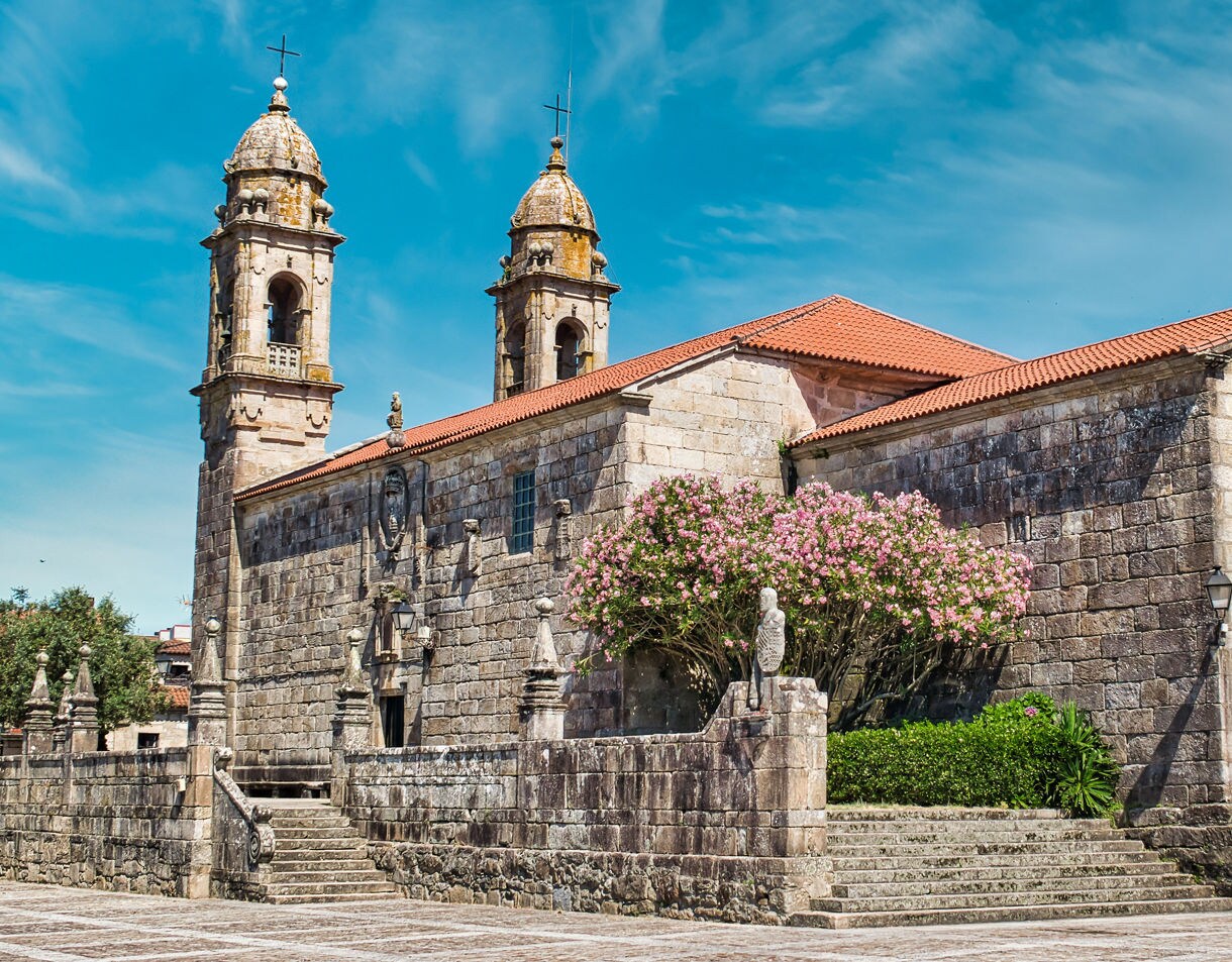  A historic stone church in Cambados, Spain, with two bell towers, red tile roofs and a blooming pink oleander tree in front, set under a bright blue sky.