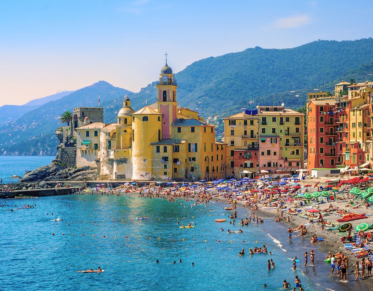 Colorful seaside buildings and beach umbrellas along the lively shoreline of Camogli, Italy, with swimmers in clear blue water.