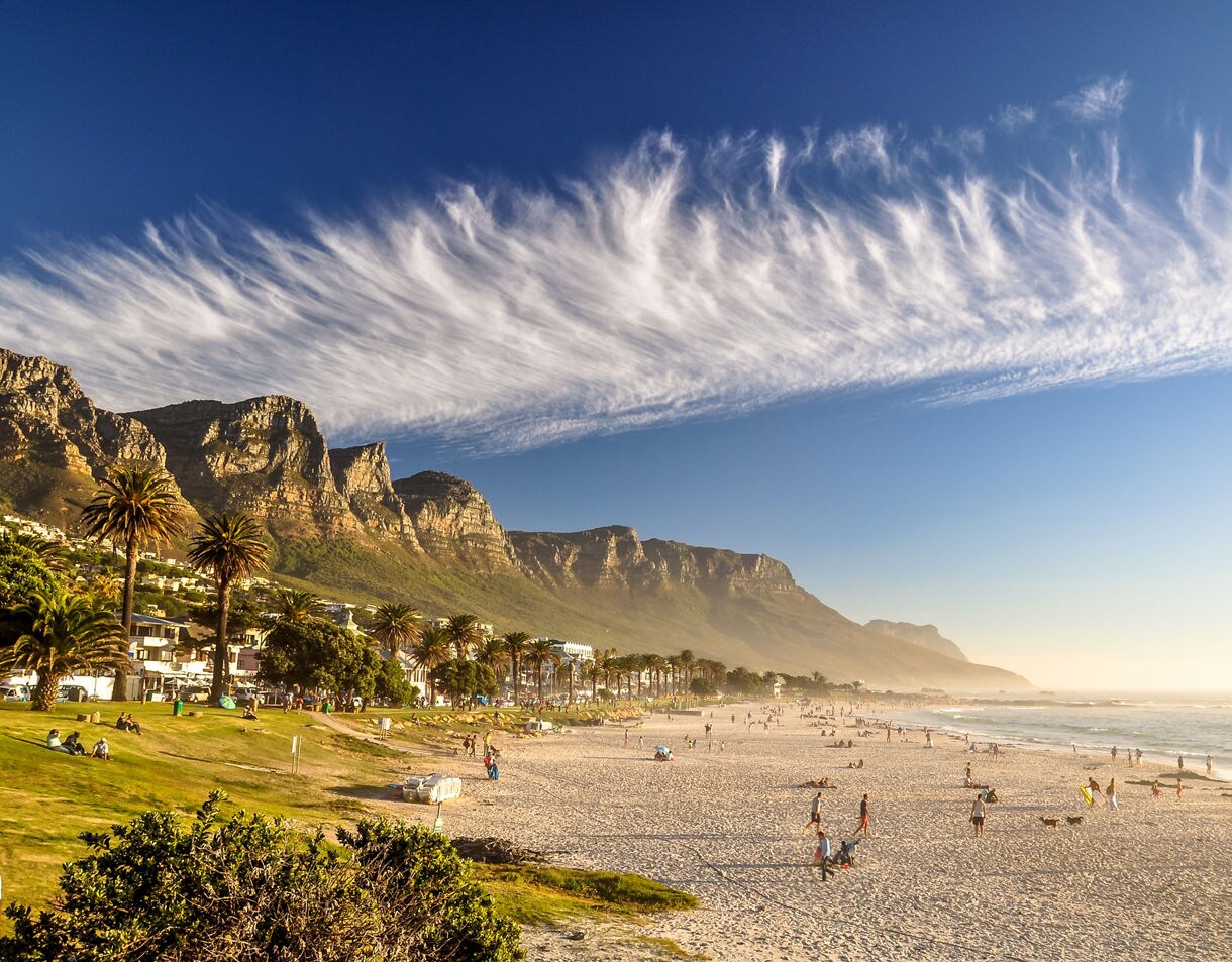 A wide sandy beach with people relaxing and playing, backed by palm trees and rugged mountains under streaked clouds.