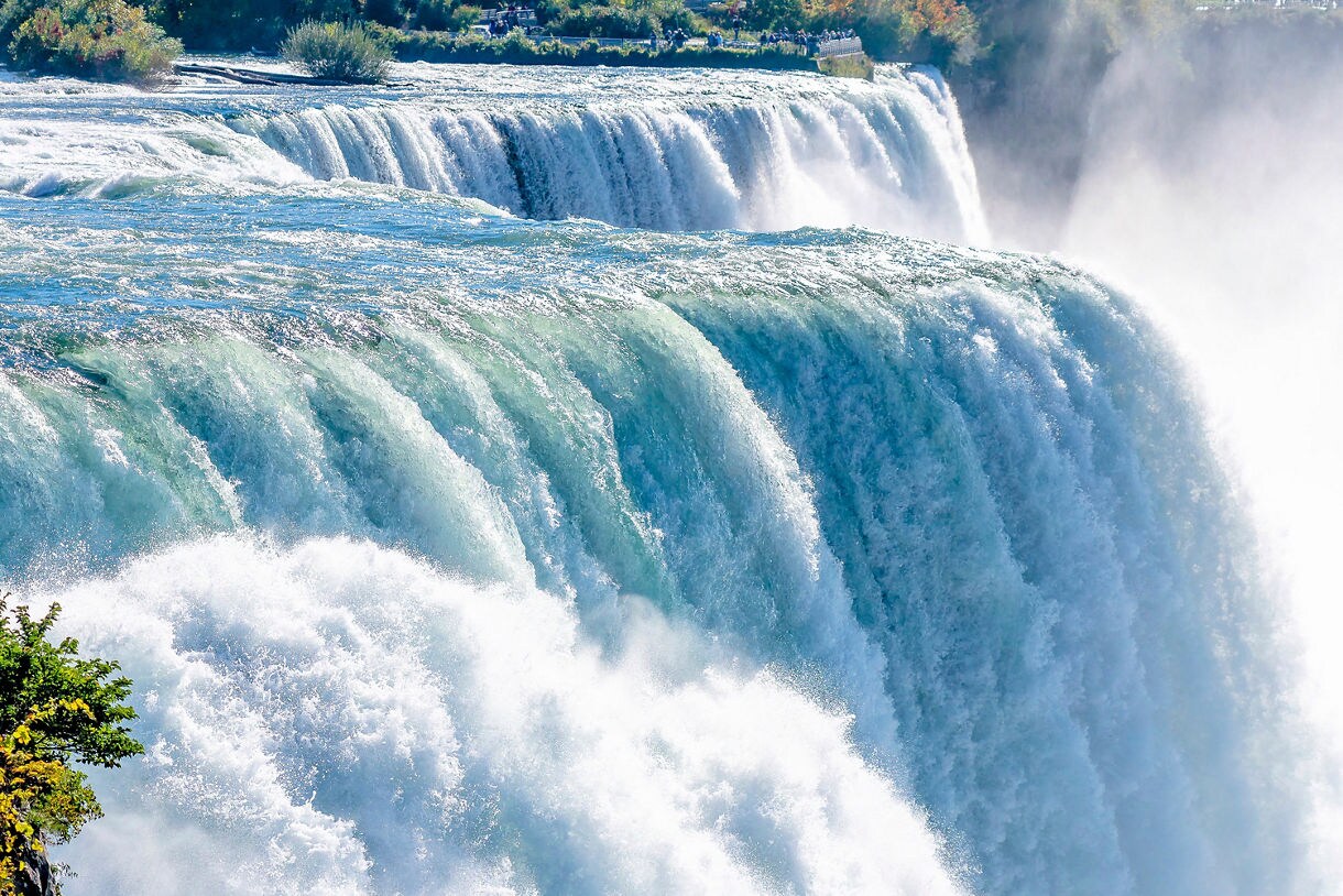 Close view of Niagara Falls with powerful turquoise water rushing over the brink and mist rising beside tree-lined cliffs.
