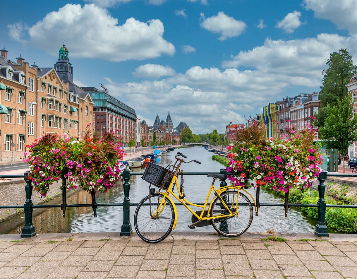 Yellow bicycle parked on a canal bridge in Amsterdam, framed by colorful flower baskets, with traditional Dutch buildings and a calm waterway stretching into the distance under a partly cloudy sky.