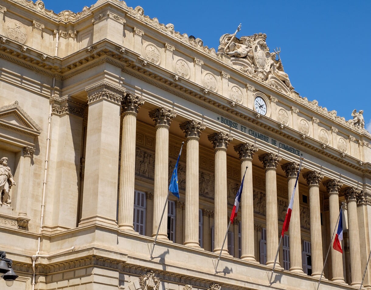 The ornate façade of the Palais de la Bourse in Marseille, featuring tall Corinthian columns, intricate carvings and French flags fluttering in front.