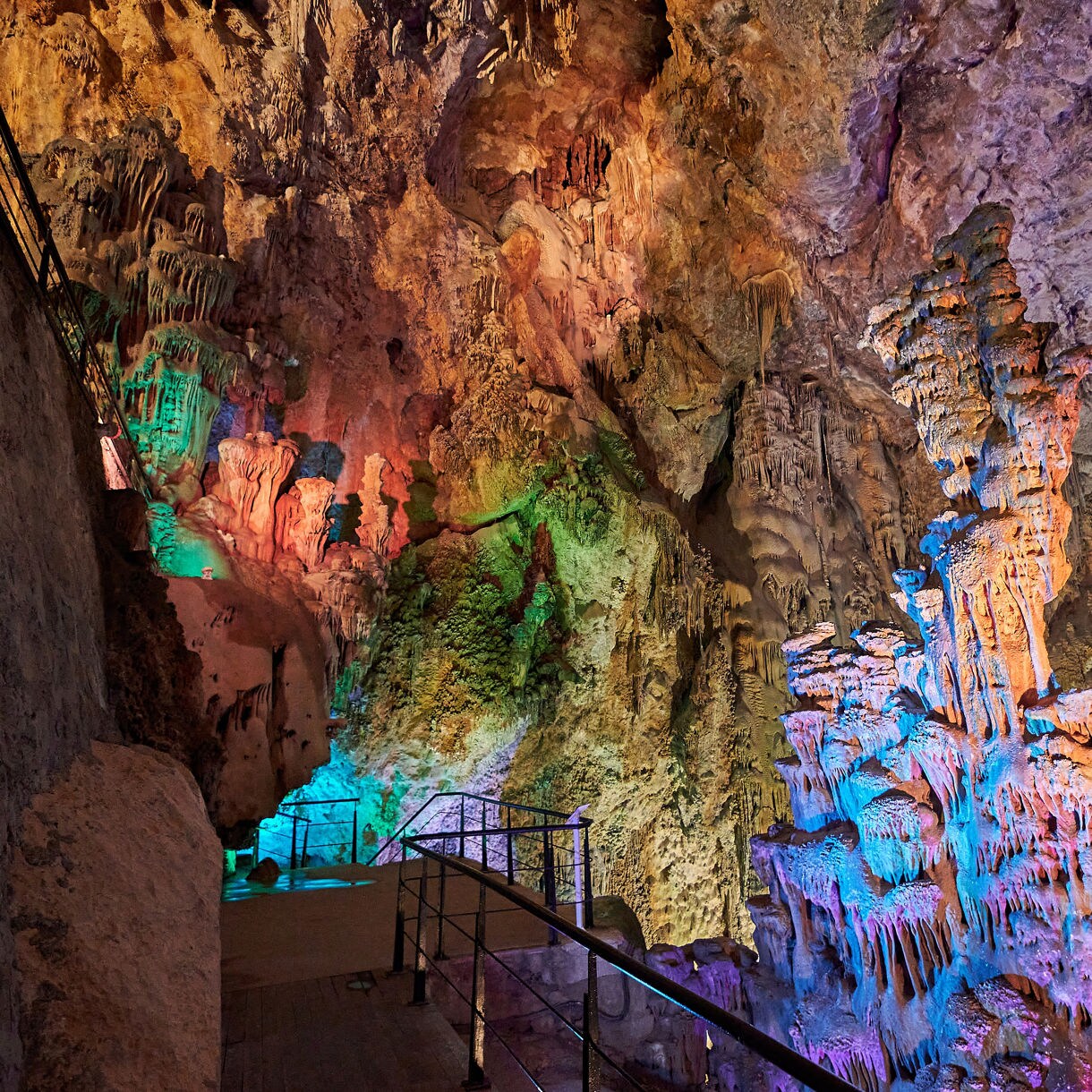 Illuminated interior of Canelobre Caves showing dramatic stalactites and stalagmites lit with vibrant blue, green and orange lights along a narrow walkway against rugged rock walls.