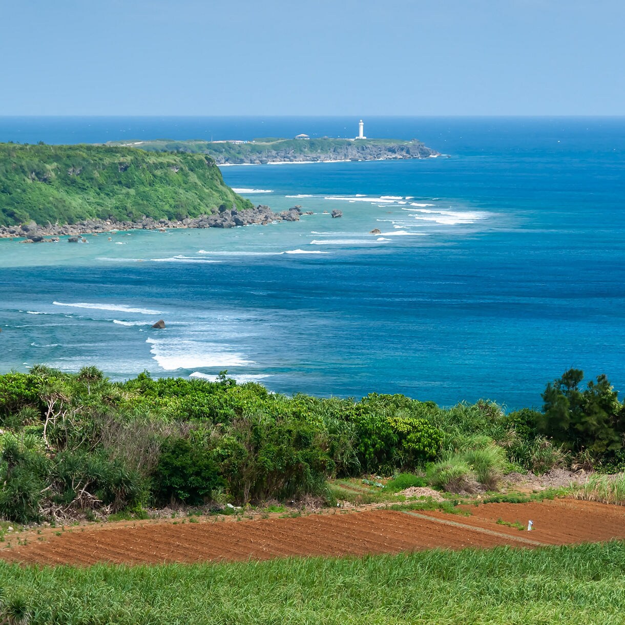  Elevated view of Cape Higashi-Hennazaki with green coastal cliffs, gentle waves rolling across bright blue water and a white lighthouse perched at the tip of the peninsula.