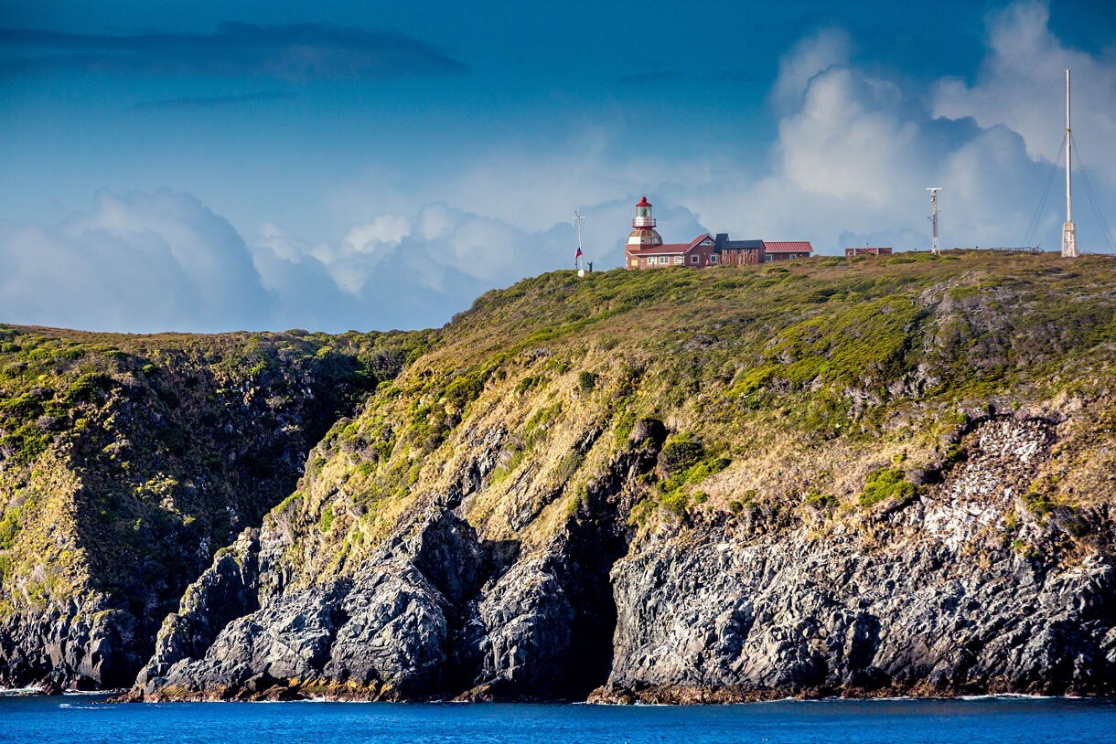 Distant lighthouse and buildings perched atop a rugged, green-covered cliff above deep blue ocean waters under a cloudy sky.