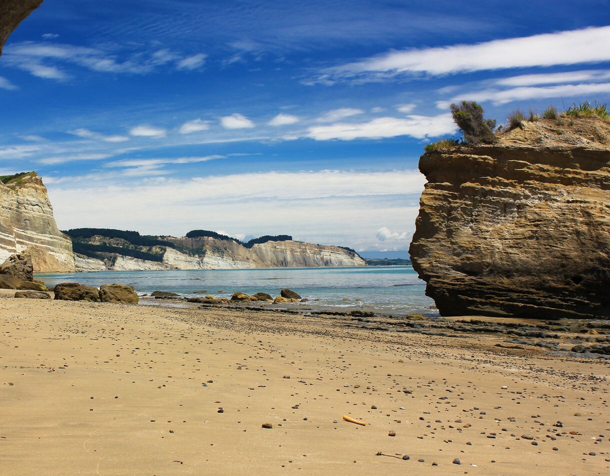 A sandy beach scattered with small stones sits below tall layered cliffs, with calm blue water and a bright sky overhead.
