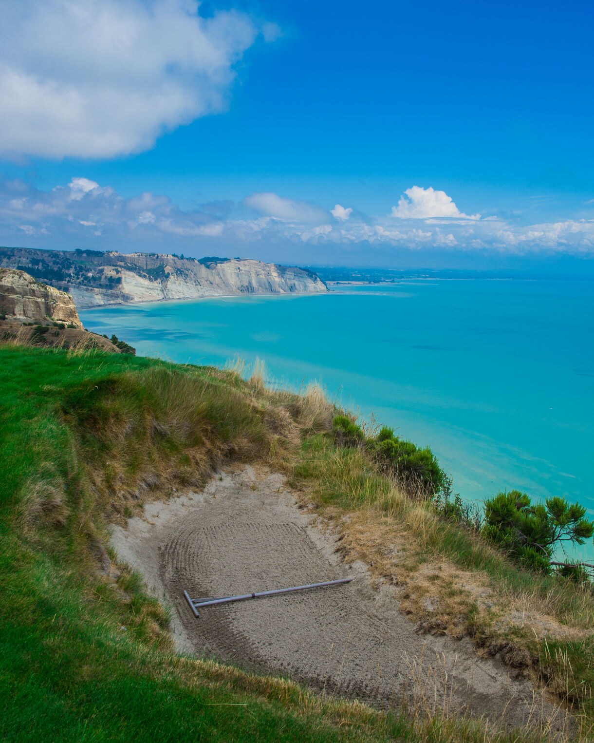 A cliffside golf course bunker overlooking bright turquoise ocean and white coastal cliffs under a vivid blue sky.