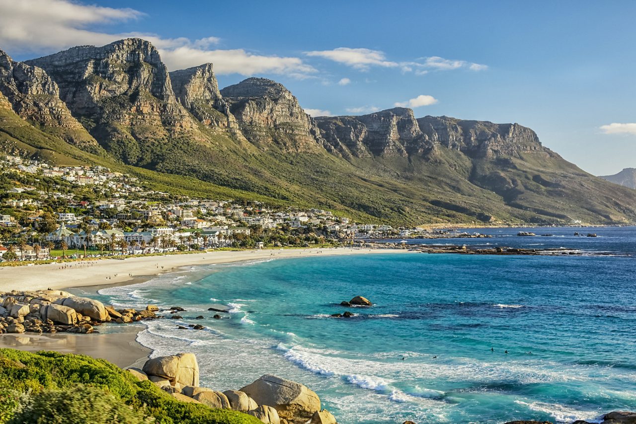 A wide sandy beach with blue ocean waves at the base of tall rocky mountains and coastal homes.