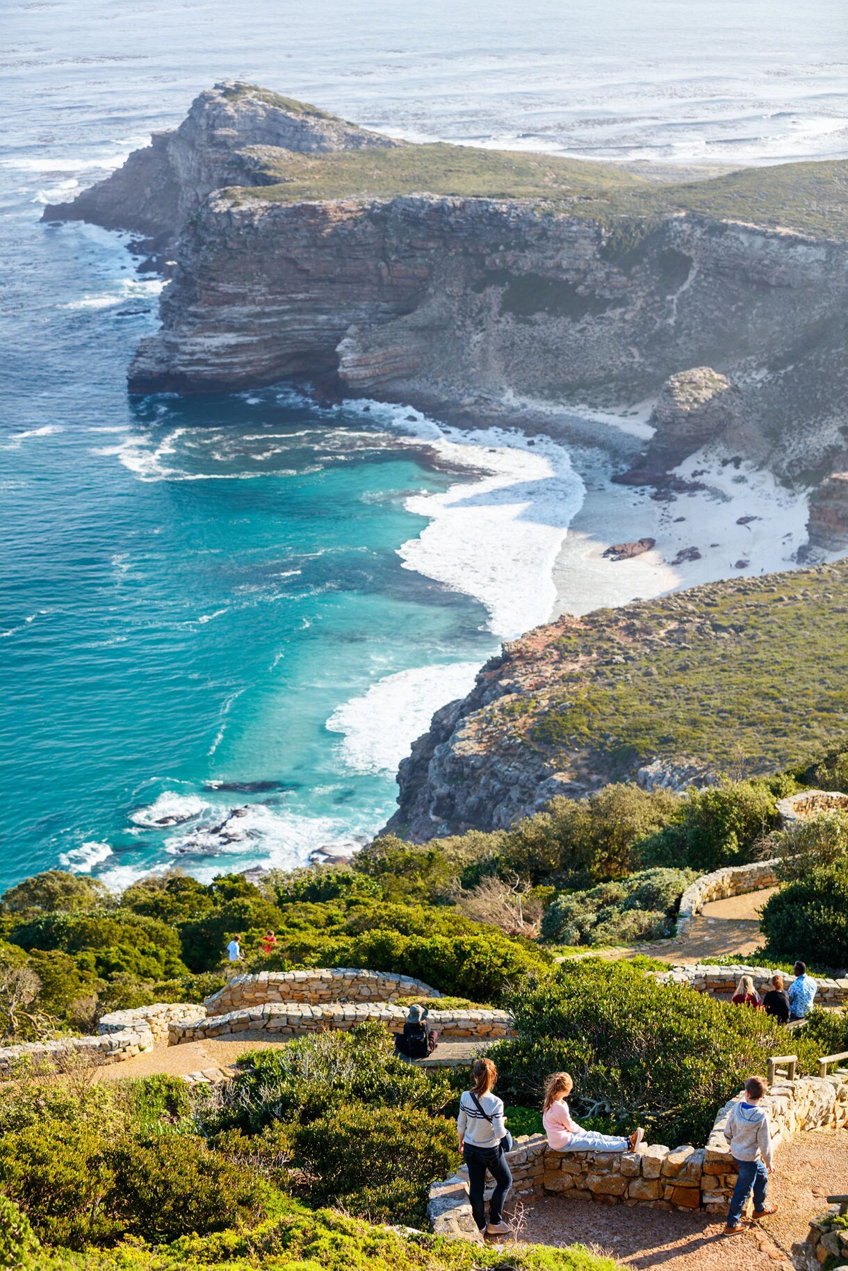 View from a cliffside lookout at the Cape of Good Hope, showing rugged coastline, turquoise waves, white-sand coves and visitors walking along stone pathways surrounded by green shrubs.