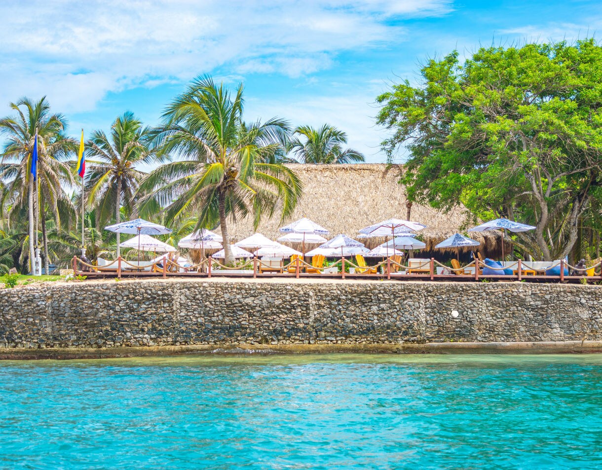 Seaside view in Cartagena with palm trees, thatched-roof cabanas, white umbrellas and lounge chairs along a stone wall beside clear blue water.