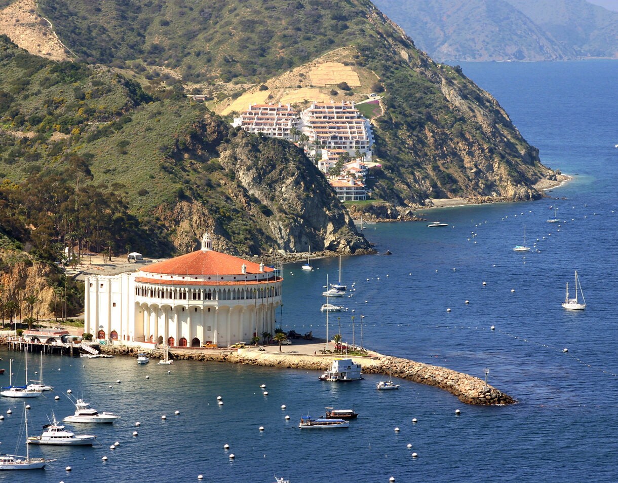 Aerial view of Catalina Island’s round, white Casino building beside a calm blue harbor dotted with moored boats and backed by rugged hills.