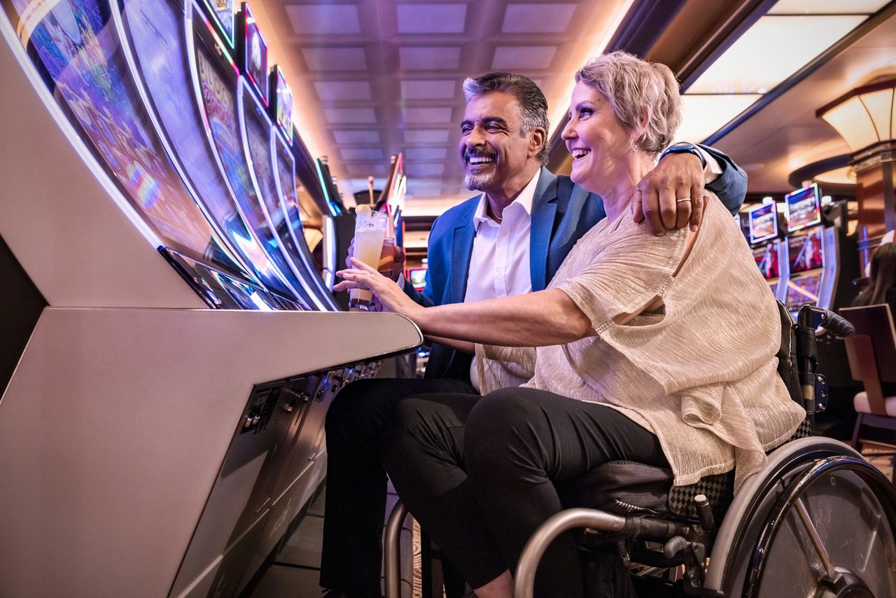 A woman in a wheelchair and her male companion laugh together while playing a slot machine in the ship's casino.