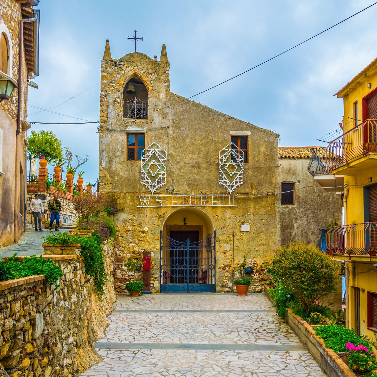 Narrow pathway leading to an old stone chapel in Castelmola, Sicily, surrounded by colorful houses, potted plants and rustic stone walls on a cloudy day.