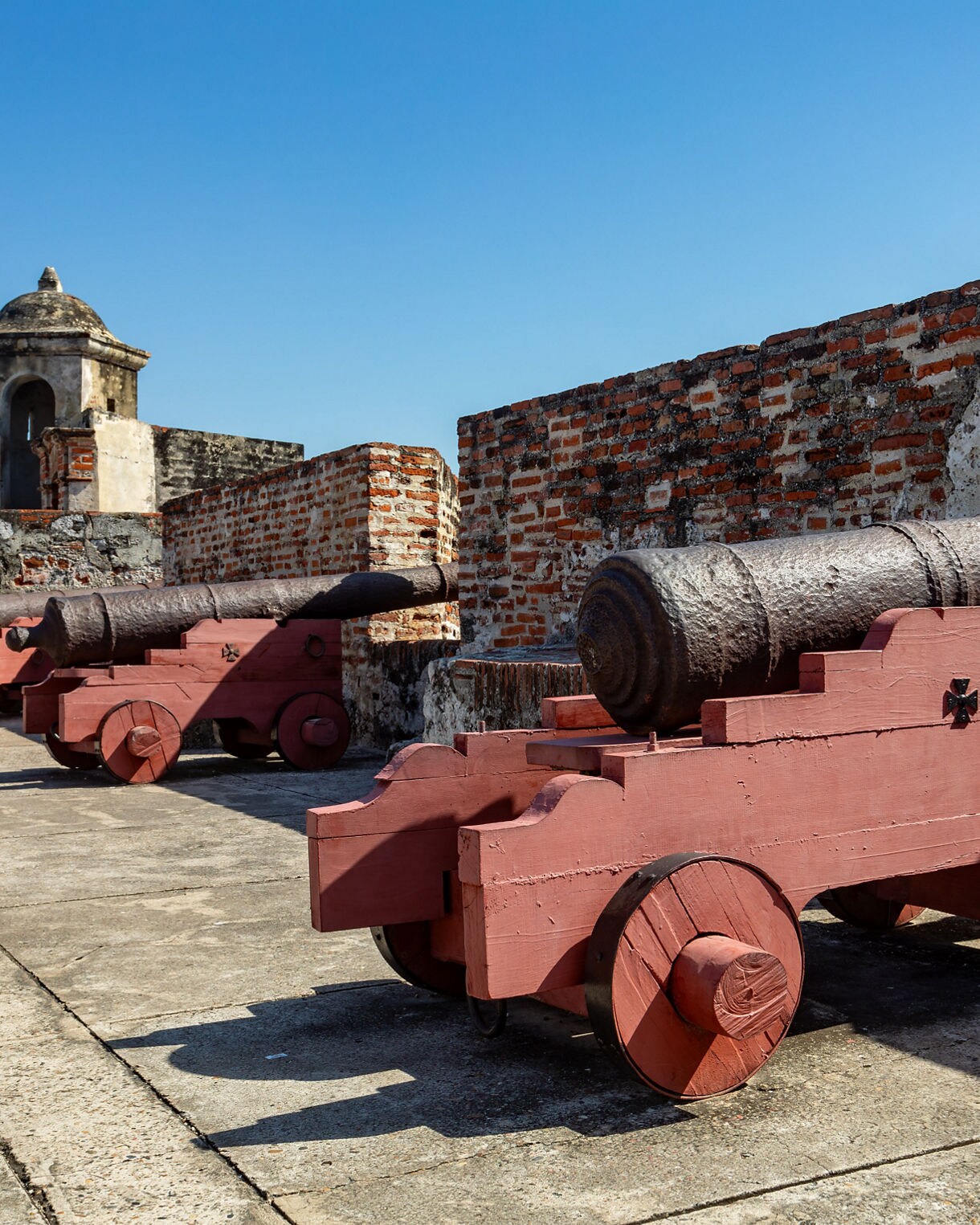Old stone fortress in Cartagena with rows of cannons mounted on red wooden carriages, set against weathered brick walls and a clear blue sky.