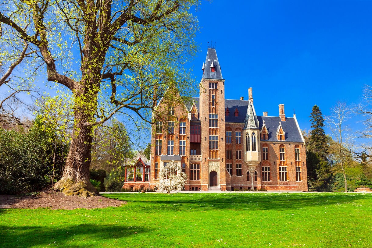 Brick Gothic Revival Loppem Castle in Belgium, surrounded by green lawns, tall trees and a bright blue sky.