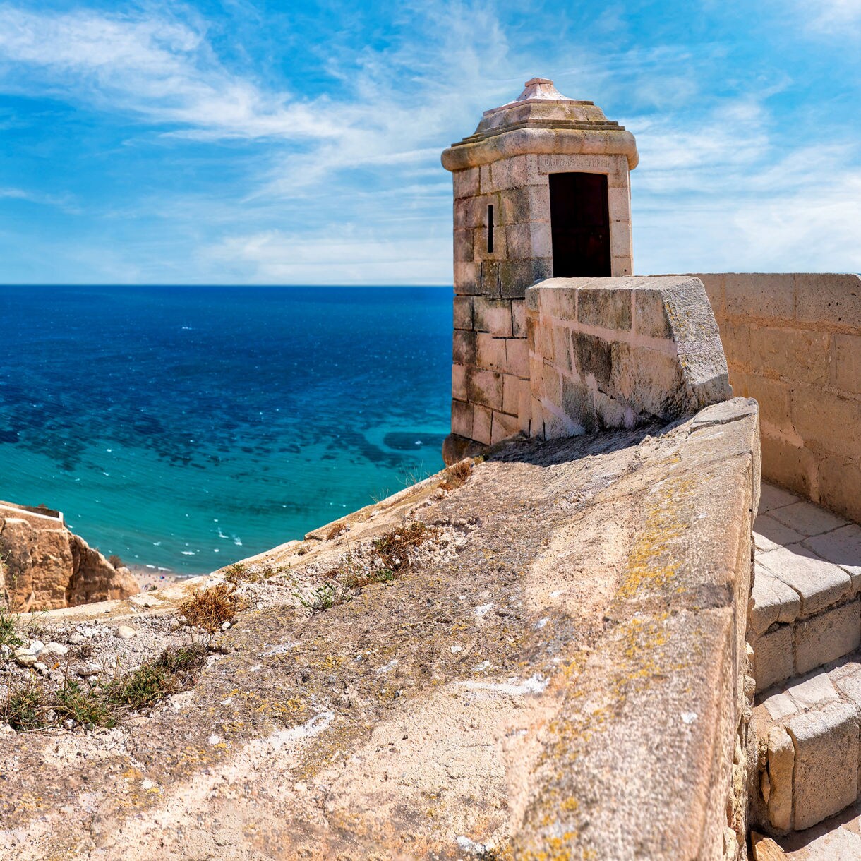 Panoramic view from Santa Bárbara Castle overlooking Alicante’s coastline, featuring stone battlements, turquoise waters and the cityscape stretching along the shore.