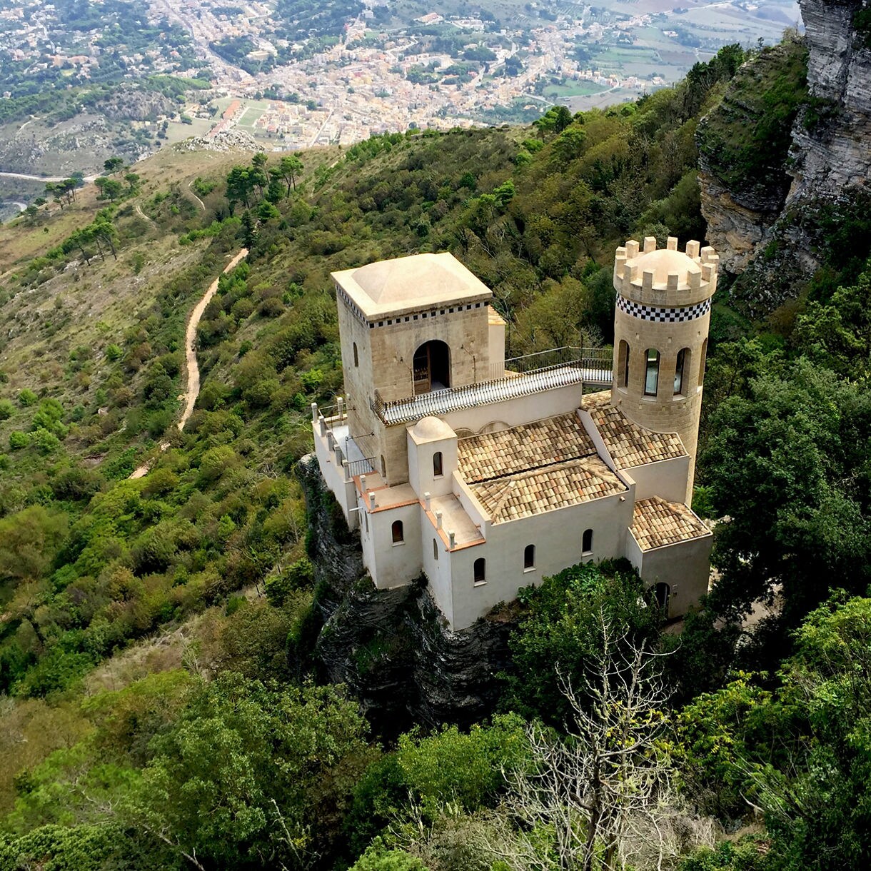 A small castle-like villa sits on a cliff surrounded by dense green forest, overlooking a wide valley and distant town.
