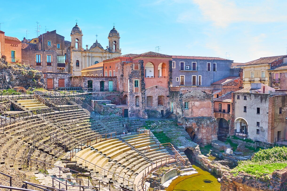 A view of Catania’s Roman theater in Sicily showing curved stone seating, partially preserved arches and surrounding historic buildings painted in warm tones under a bright blue sky.