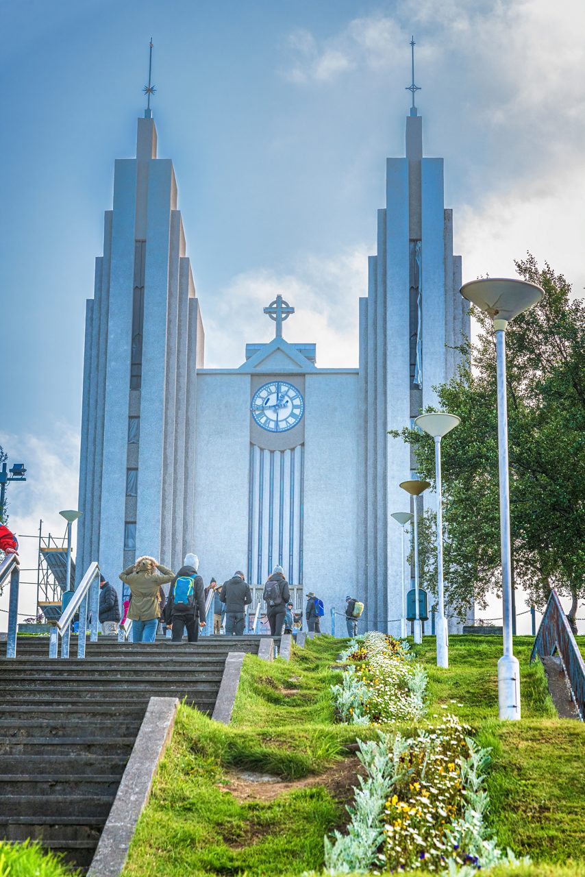 Akureyri Church in Iceland, a modernist gray concrete building with twin towers and a central clock, viewed from a staircase lined with flowers and visitors walking up.