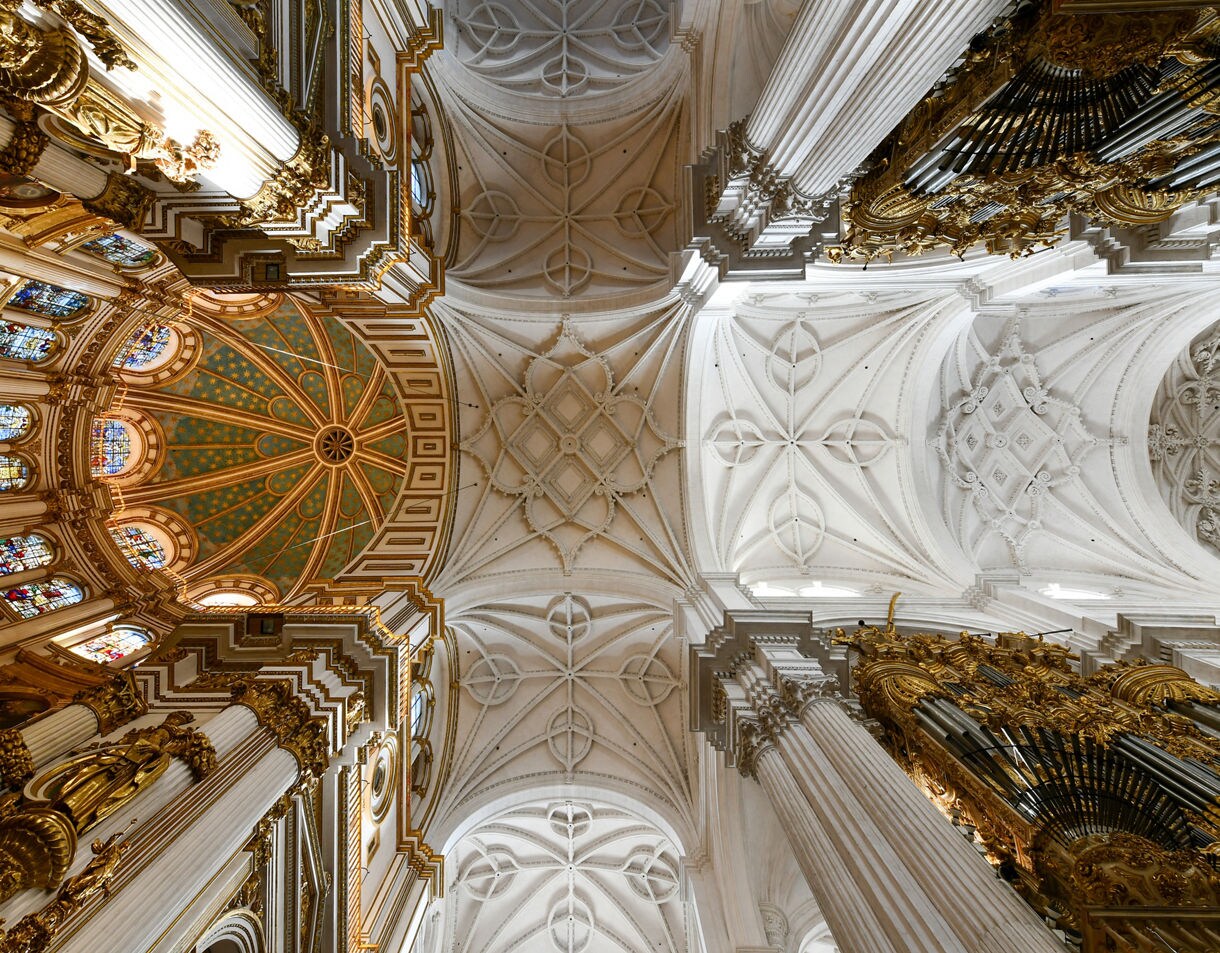 Ornate interior ceiling of Granada Cathedral, featuring white vaulted arches, intricate stonework, gold details and stained-glass windows.