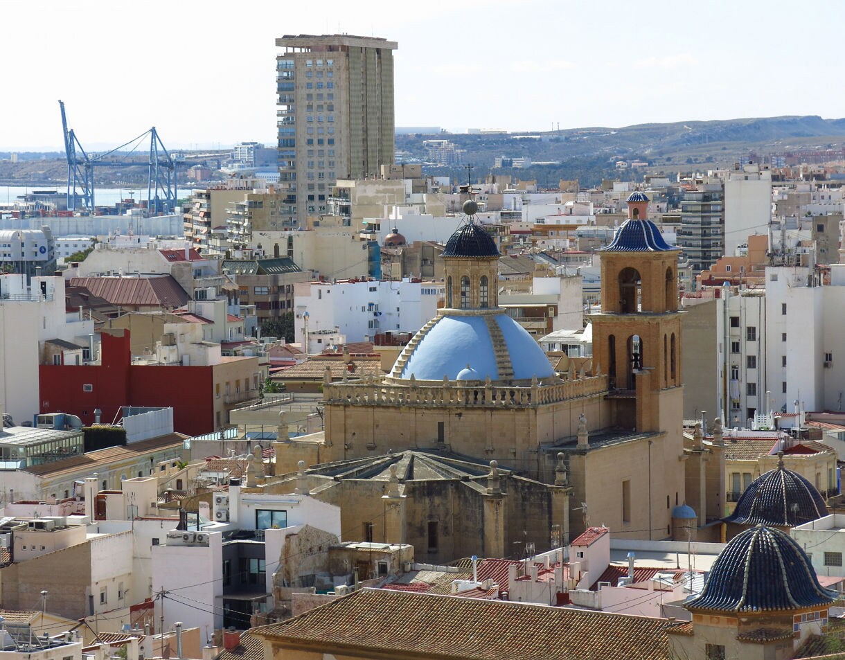 Elevated view of the Cathedral of Saint Nicholas in Alicante with its blue domes and bell tower surrounded by dense city buildings, with distant hills and a port crane visible in the background.