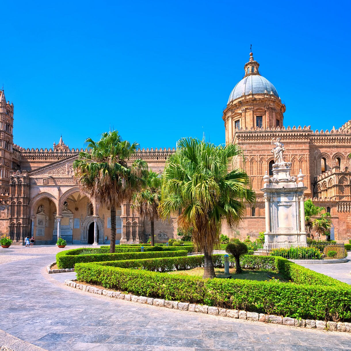 The Cathedral of Palermo with its ornate towers, domed roof and detailed stone façade, surrounded by palm trees and manicured garden paths under a bright blue sky.