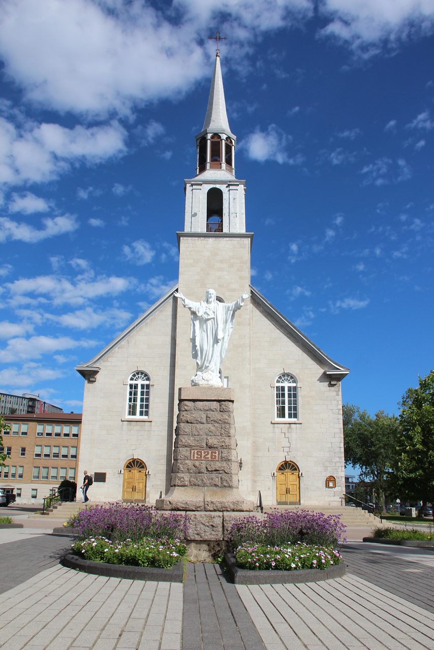 Stone Catholic church with a tall white steeple and a large statue of Christ on a stone pedestal dated 1926, framed by flower beds and a blue sky with scattered clouds.