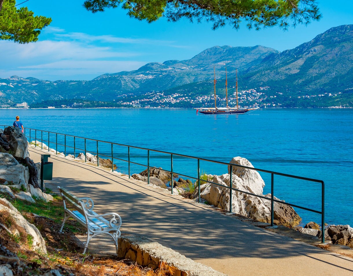 Seaside promenade in Cavtat, Croatia, with a walking path, benches, pine trees and a sailboat anchored on calm blue waters against mountain backdrops.
