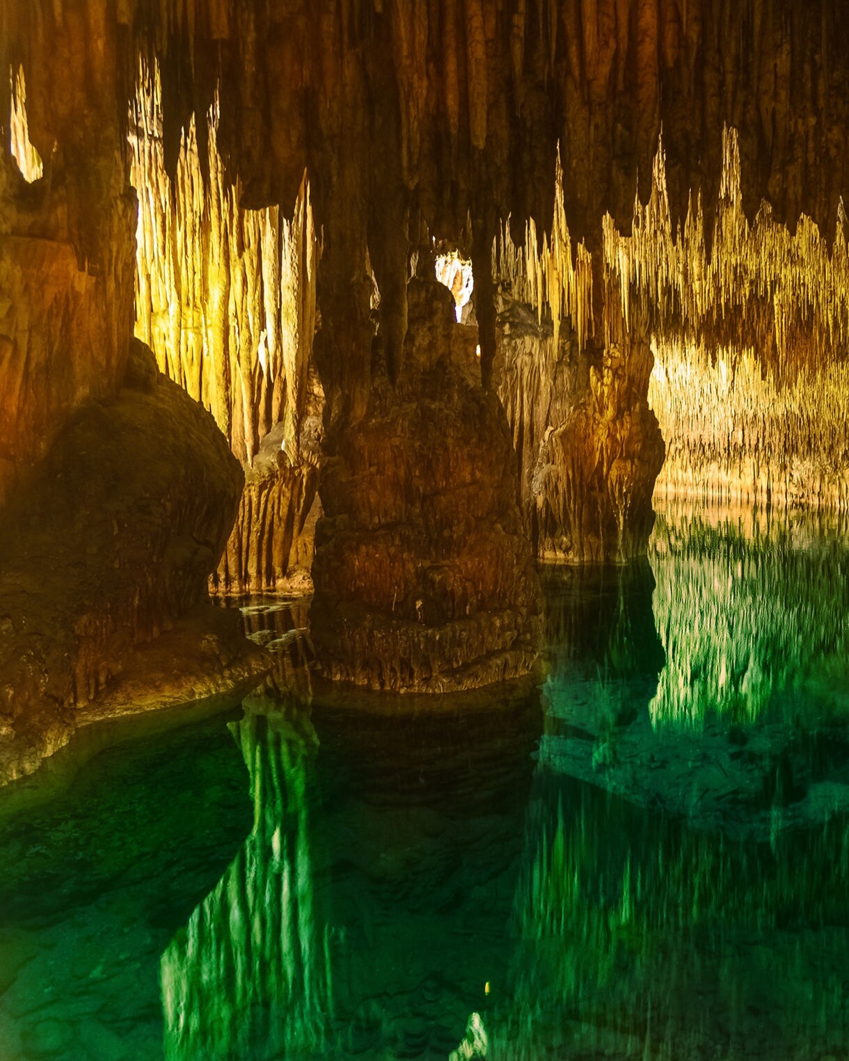 Illuminated view of the Caves of Drach in Mallorca, showing jagged stalactites and greenish underground water with mirrored reflections.
