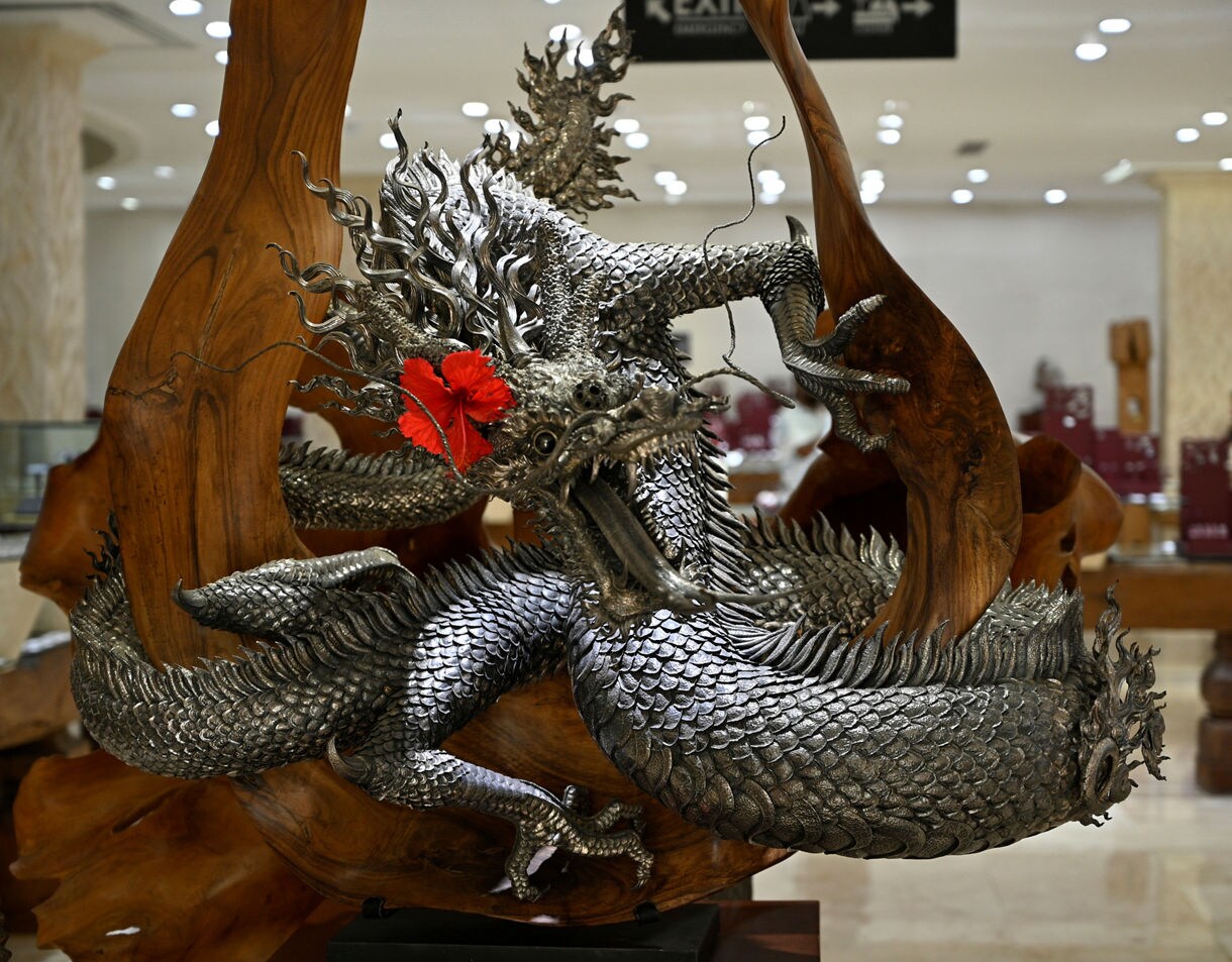 Intricate silver dragon sculpture wrapped around a carved wooden base, featuring detailed scales, flowing whiskers and a bright red hibiscus flower tucked near its head, displayed in a well-lit showroom.