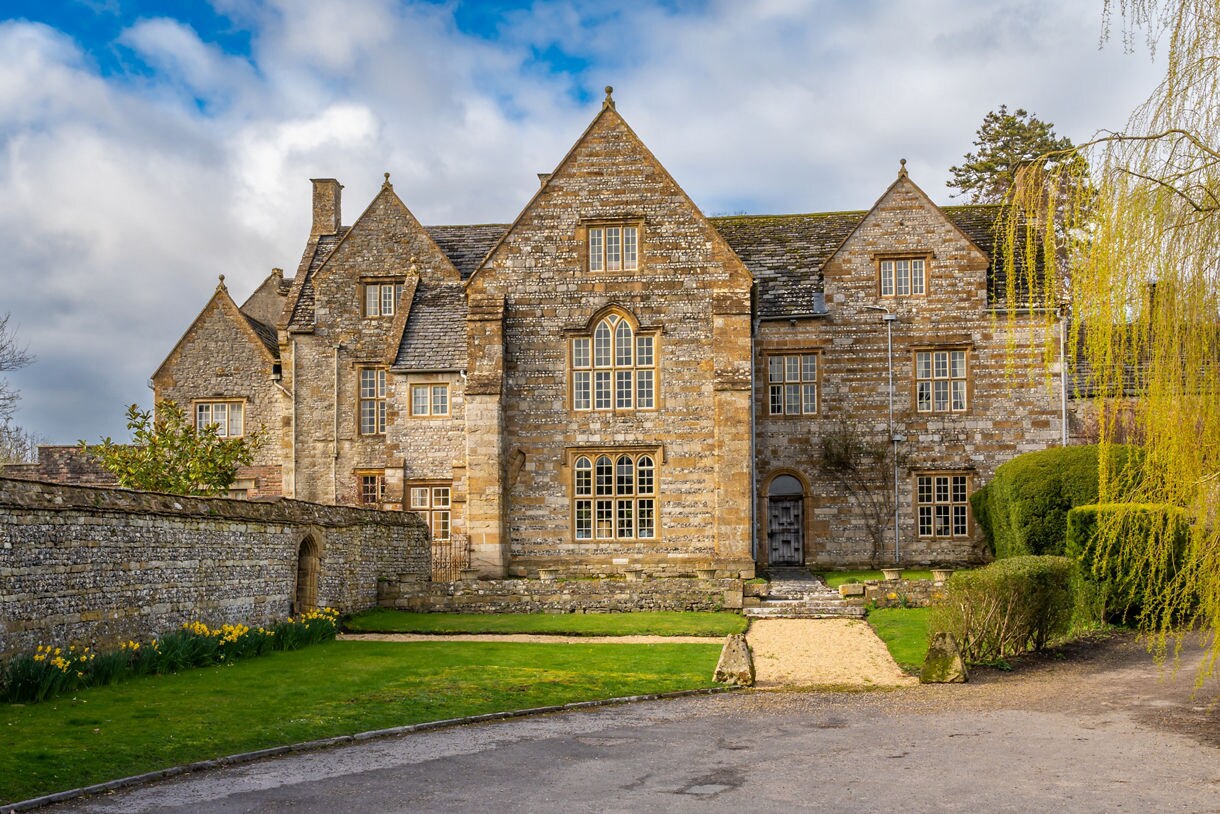 Historic stone building in Cerne Abbas, Dorset, featuring gabled roofs, mullioned windows and a walled garden with spring daffodils under a partly cloudy sky.