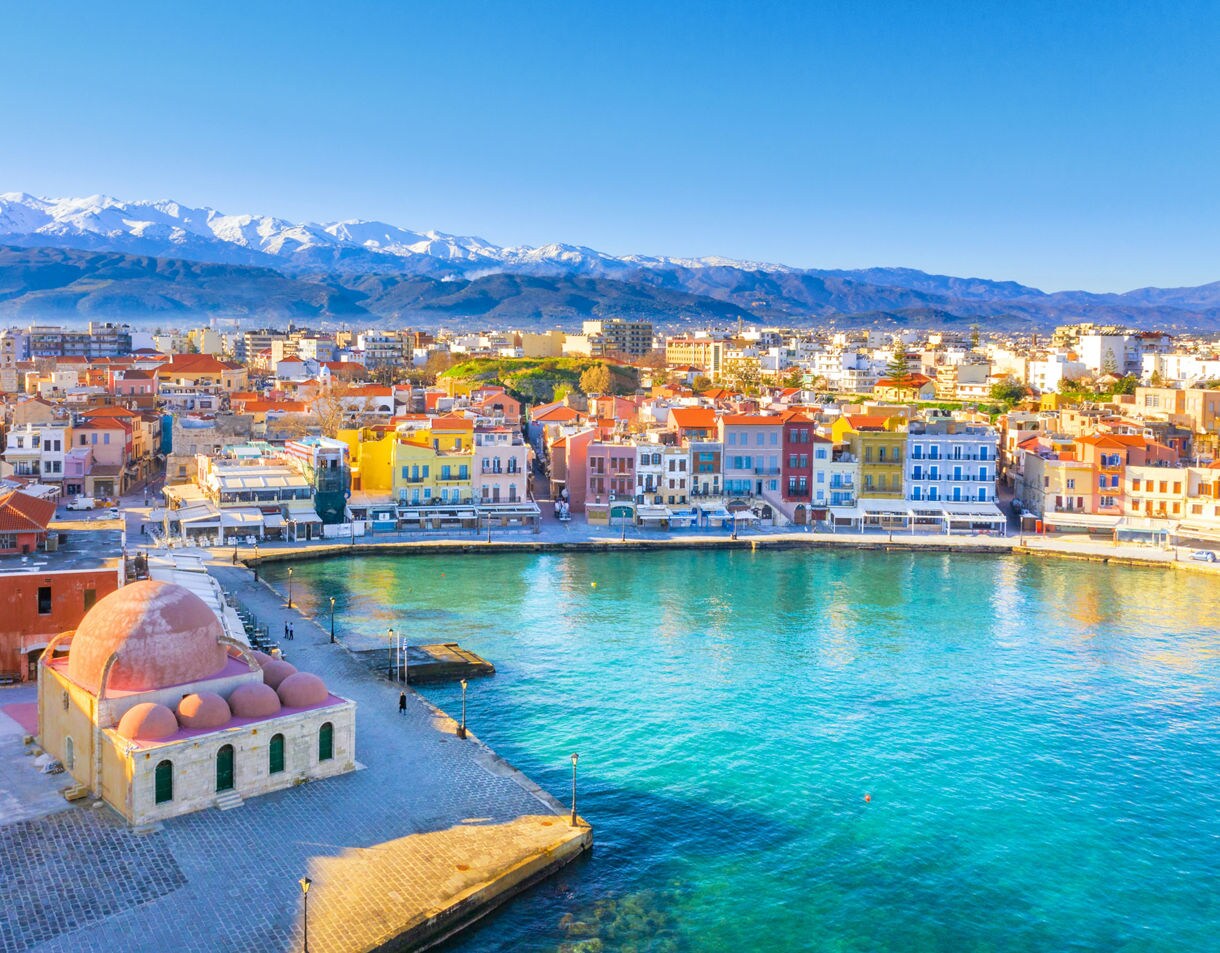 Aerial view of Chania’s Old Venetian Harbor in Crete, Greece, with colorful waterfront buildings, clear turquoise water and snowcapped mountains in the distance.